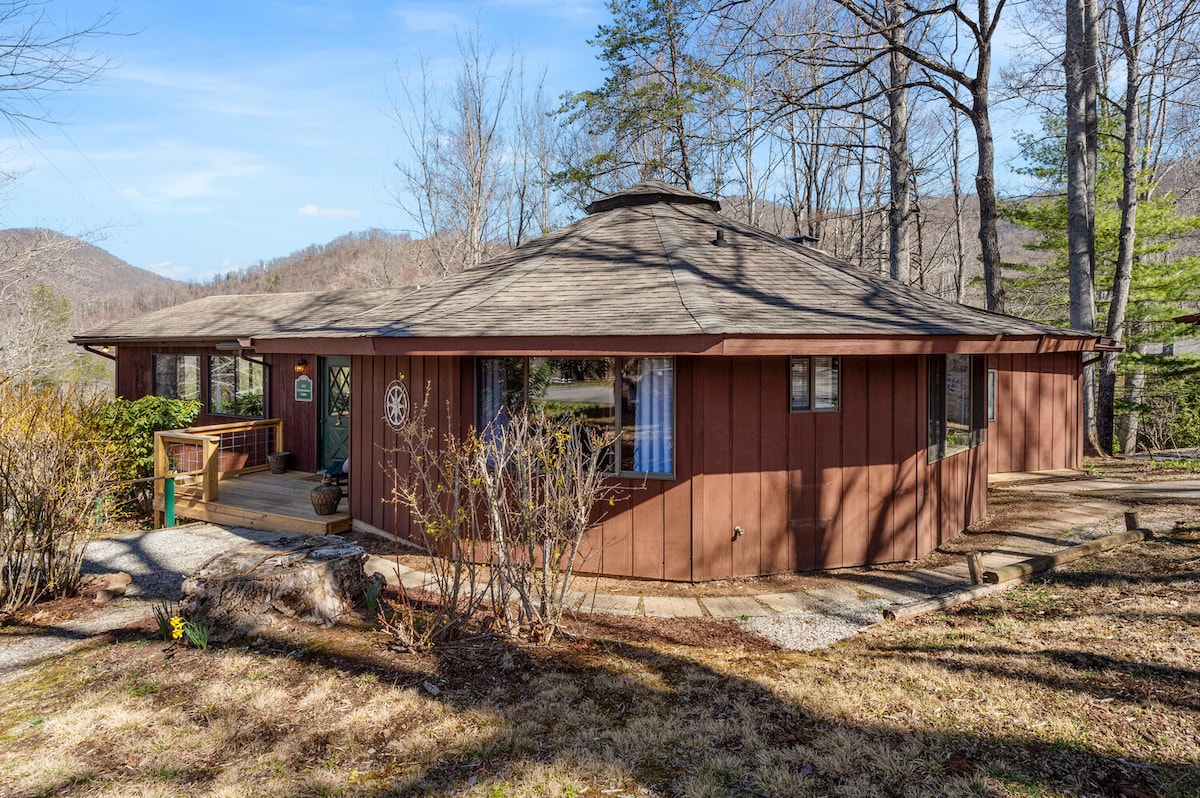 A round, single-story cabin structure is surrounded by trees and gentle slopes. Wide windows allow natural light to fill the space, and a wooden porch is visible at the entrance. Light brown siding contrasts with the landscape, creating a harmonious appearance.