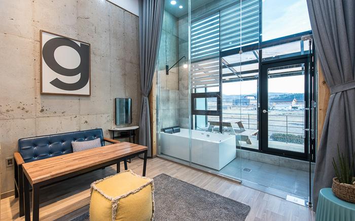 The interior space features a modern seating area with a blue sofa and a wooden table. Large glass doors provide views of the outside, with natural light illuminating the room. Relaxing decor elements like a patterned rug and a potted plant enhance the ambiance.