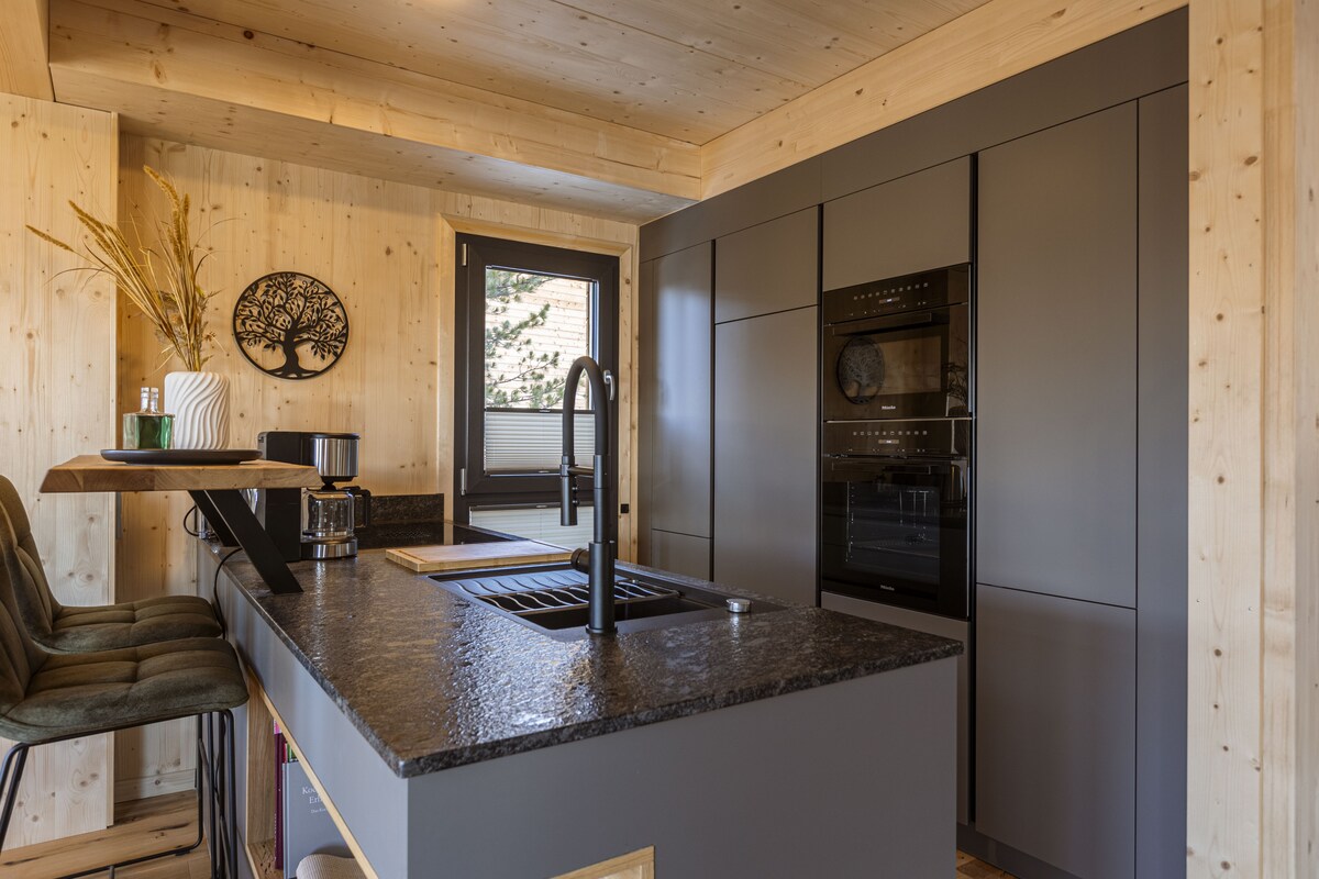 The kitchen features modern cabinetry and a sleek black countertop. An island with seating provides a casual dining option. Built-in appliances include an oven and coffee machine, while natural light enters through the window, offering a view of the surrounding landscape.