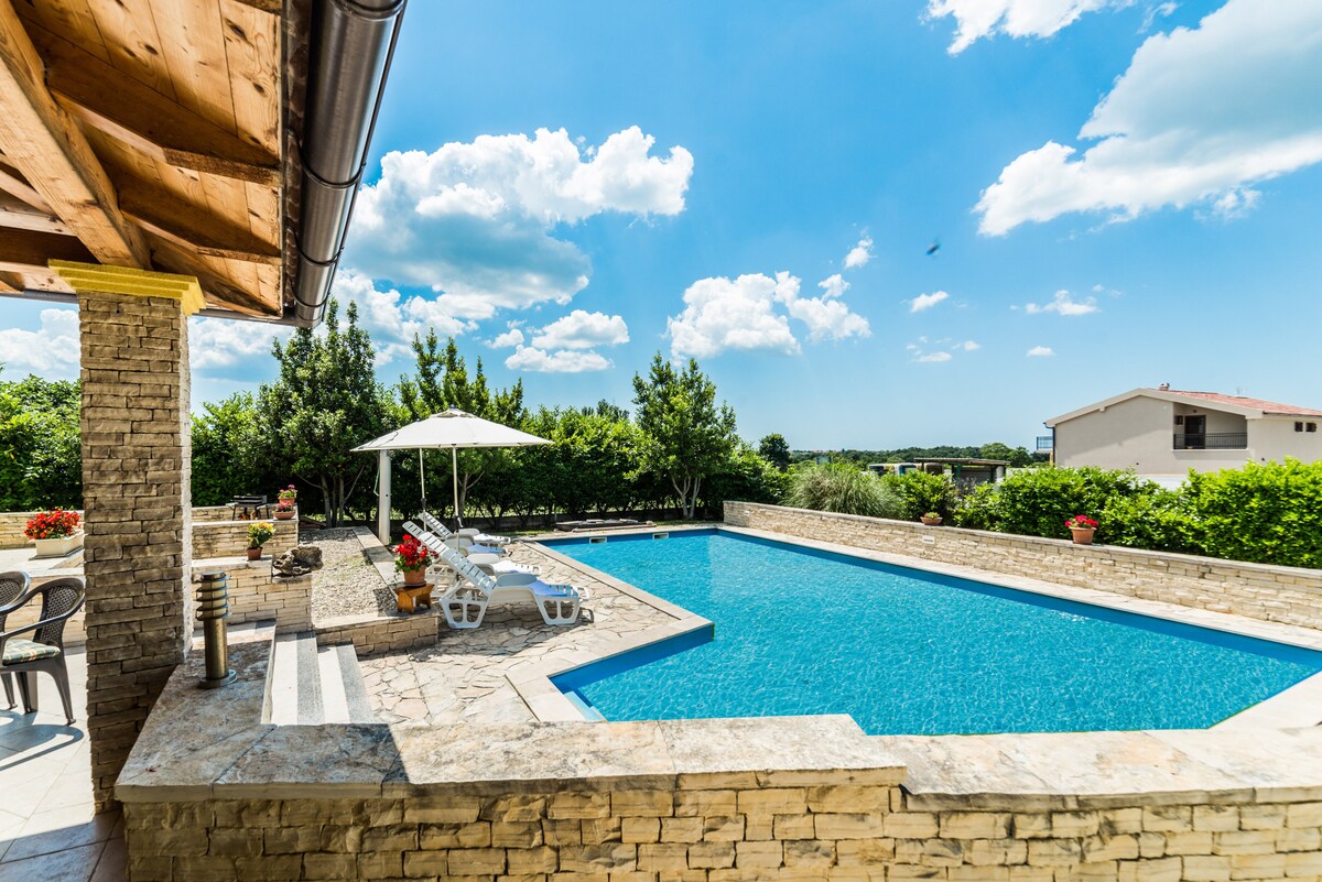 A large outdoor swimming pool glistens in the sunlight, surrounded by a stone patio. Sun loungers are arranged beside the pool, while a shaded area with an umbrella provides relief from the sun. Lush green foliage frames the scene, enhancing the tranquil atmosphere.