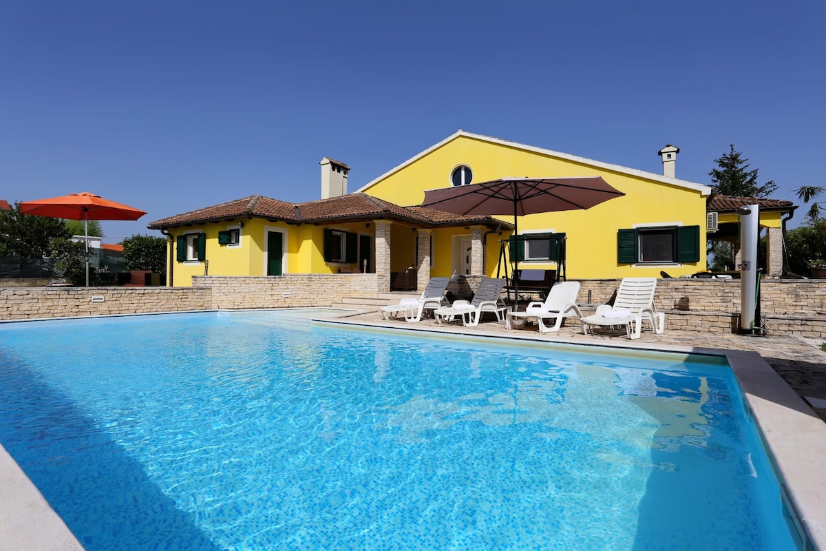 The exterior view showcases a spacious yellow villa beside a refreshing swimming pool. Sun loungers are arranged around the pool, with an orange umbrella providing shade. Lush greenery surrounds the property, enhancing the tranquil atmosphere.