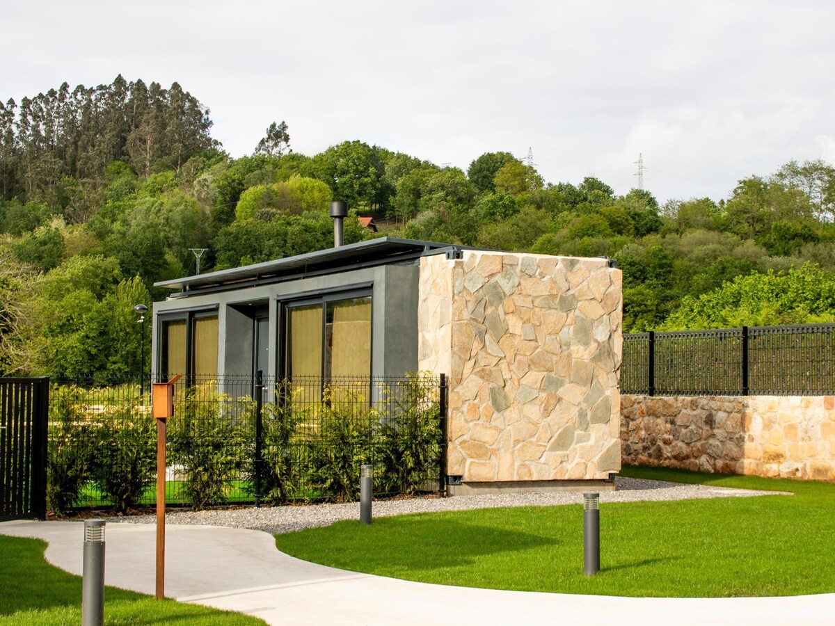 A modern bungalow features a stone and glass exterior, surrounded by lush greenery. Smooth pathways meander through the manicured lawn, leading to the entrance. Large windows reflect the natural light, offering views of the serene landscape, while a wrought iron fence outlines the property.