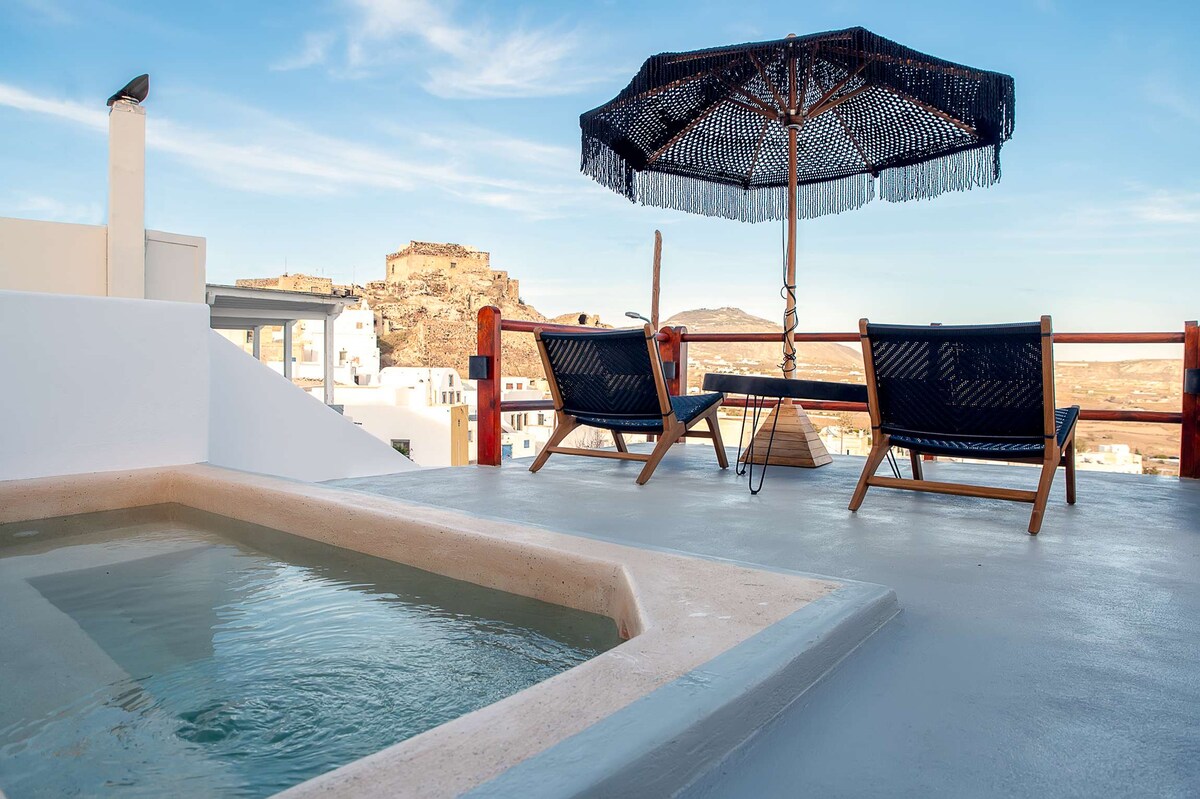 A private terrace features a small, inviting hot tub surrounded by a smooth, white surface. Two wooden lounge chairs are positioned under a decorative umbrella, providing a comfortable space to enjoy the scenic views of the surrounding landscape.