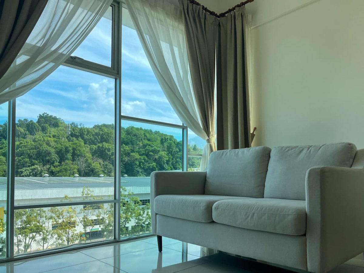 A light-filled living area features a soft two-seater sofa positioned near large windows. Flowing curtains frame the window, providing a view of lush greenery outside. The space is designed for relaxation and comfort.