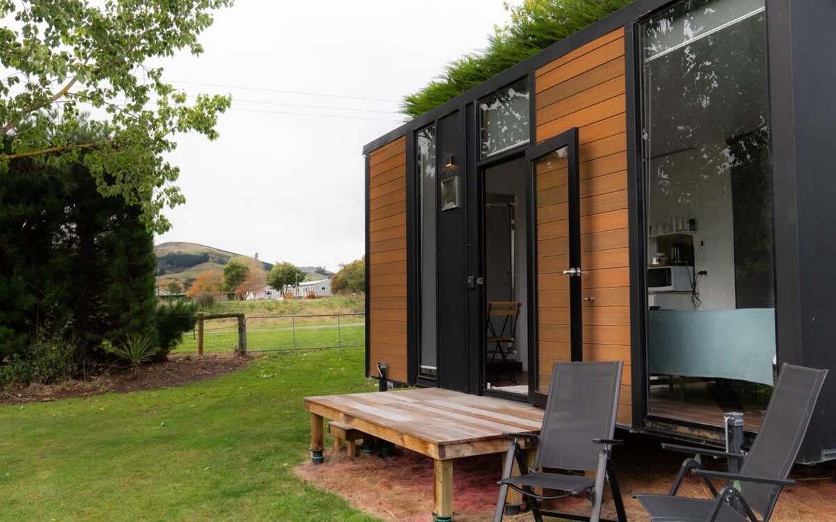 A modern tiny home is set against a backdrop of rolling hills. The exterior features warm wooden panels and large windows. Outdoor seating is provided with two black chairs positioned on a wooden deck, inviting relaxation in the serene landscape.