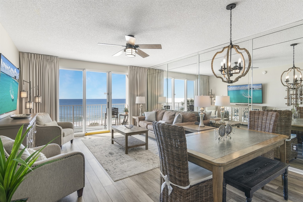 A bright living area features a comfortable seating arrangement in neutral tones, complemented by a large wooden dining table. Floor-to-ceiling mirrors reflect views of the ocean through glass doors, which open to a spacious balcony, enhancing the coastal ambiance.