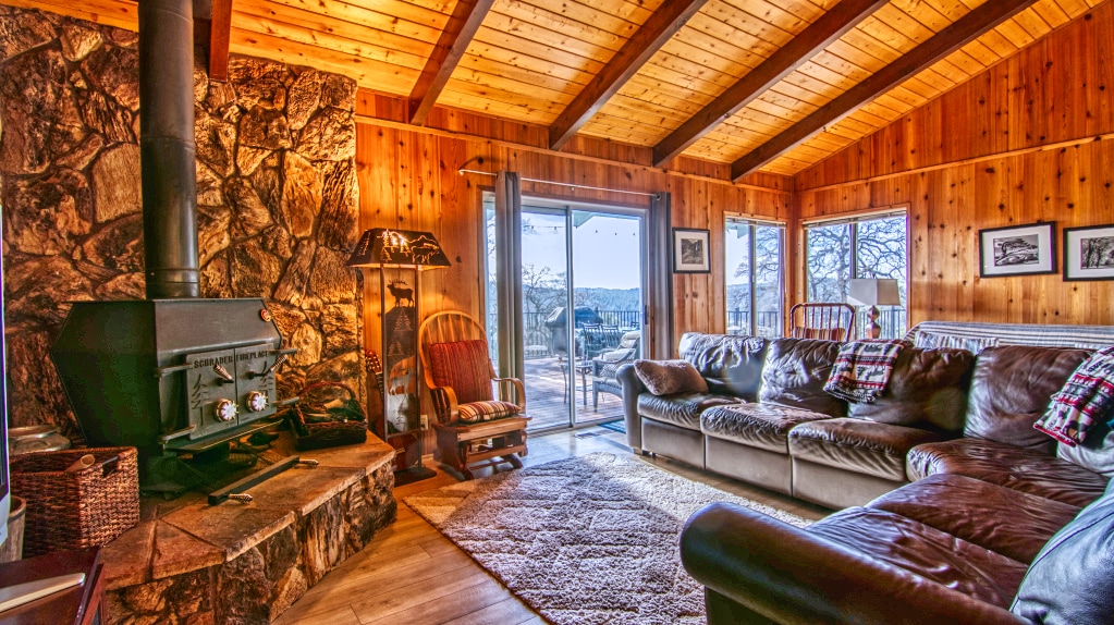 The great room features a cozy seating area with a leather sectional and wooden beams overhead. A stone fireplace stands prominently, while large windows provide natural light and views of the surroundings. A door leads to an outdoor space, enhancing the connection to nature.