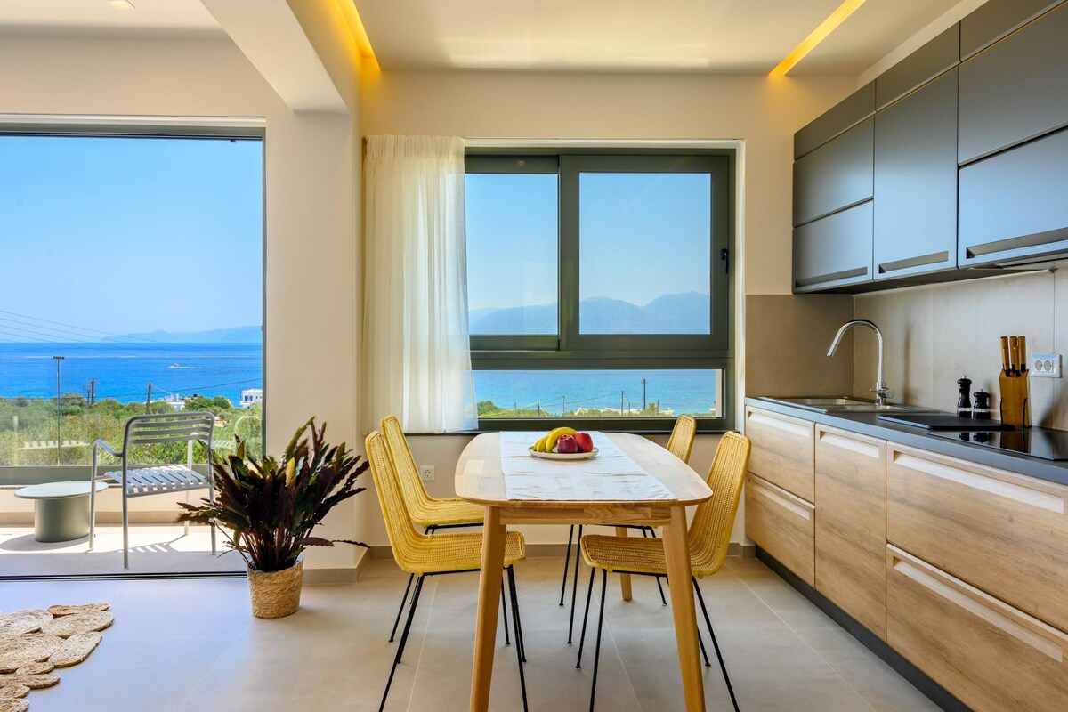 A modern kitchen space is highlighted by a wooden dining table surrounded by four wicker chairs. Large windows offer a view of the sea, while a fully equipped kitchen with contemporary cabinetry and appliances is positioned nearby. Natural light brightens the inviting space.