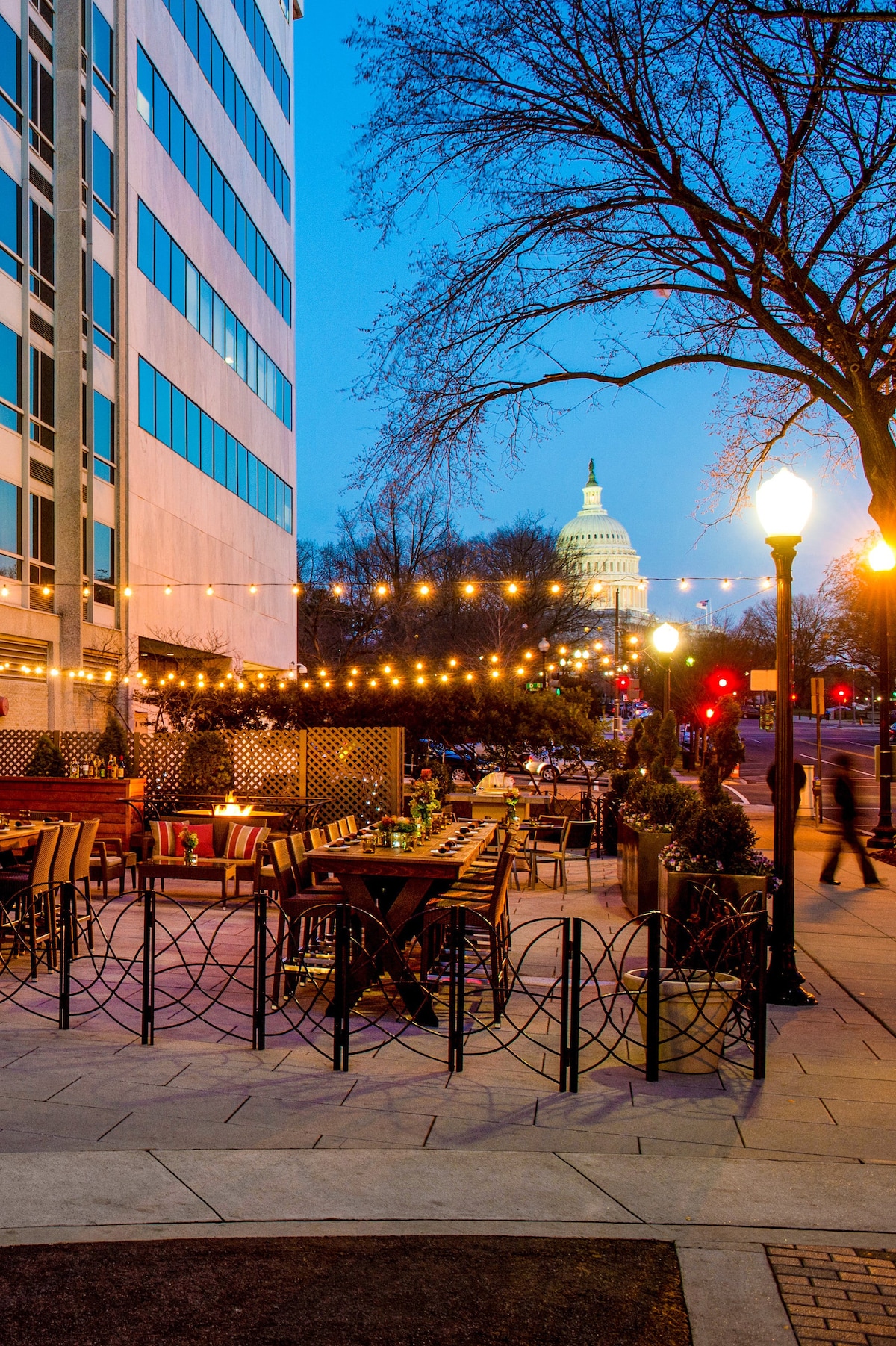 An outdoor dining area is visible, featuring wooden tables and cozy seating under string lights, creating a welcoming ambiance. In the background, the dome of the Capitol building is illuminated against a twilight sky, enhancing the lively city atmosphere.