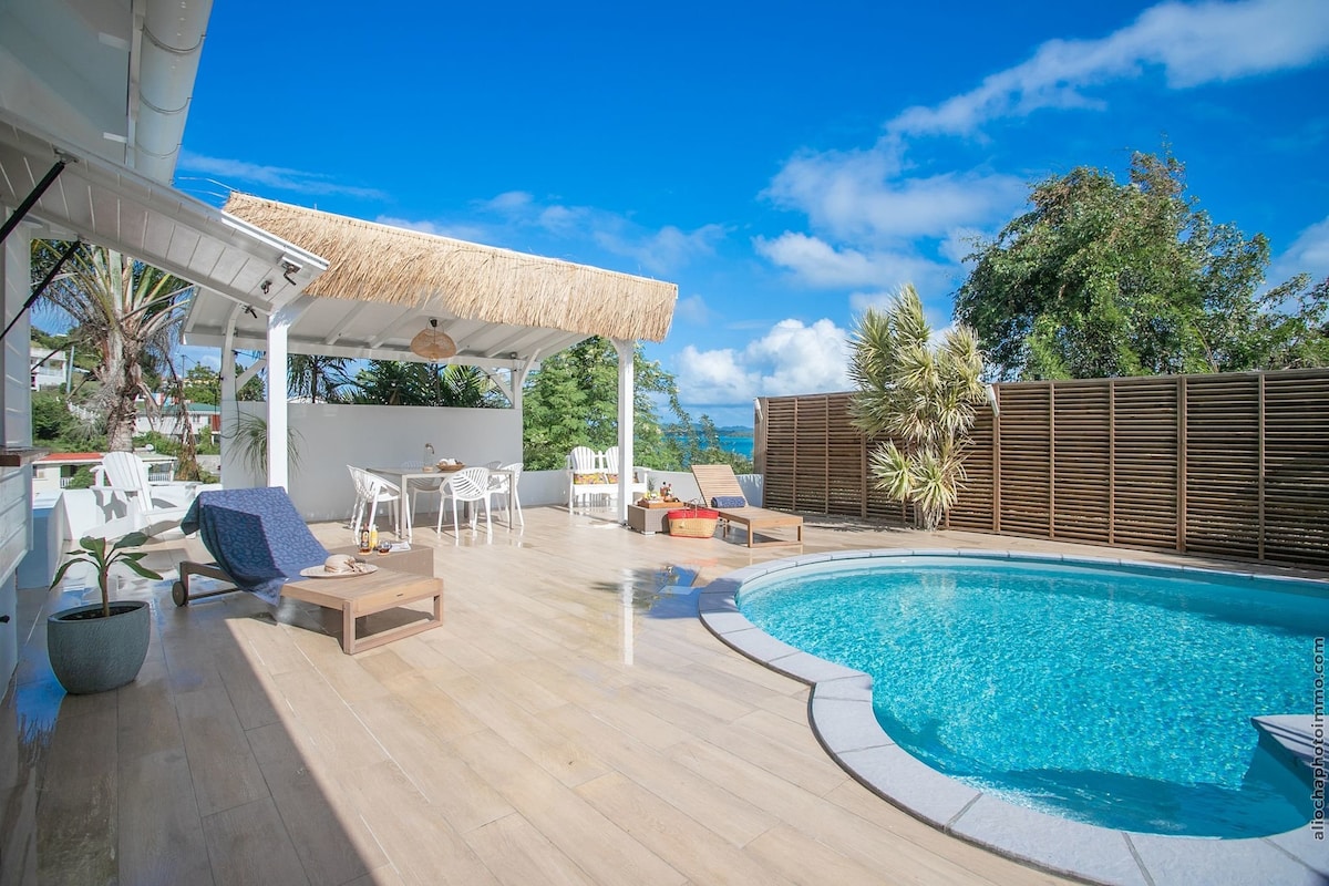 An outdoor terrace showcases a private swimming pool surrounded by wooden decking. A sheltered seating area features a thatched roof, while lounge chairs and a small table offer space for relaxation. Lush greenery and a clear blue sky enhance the inviting atmosphere.