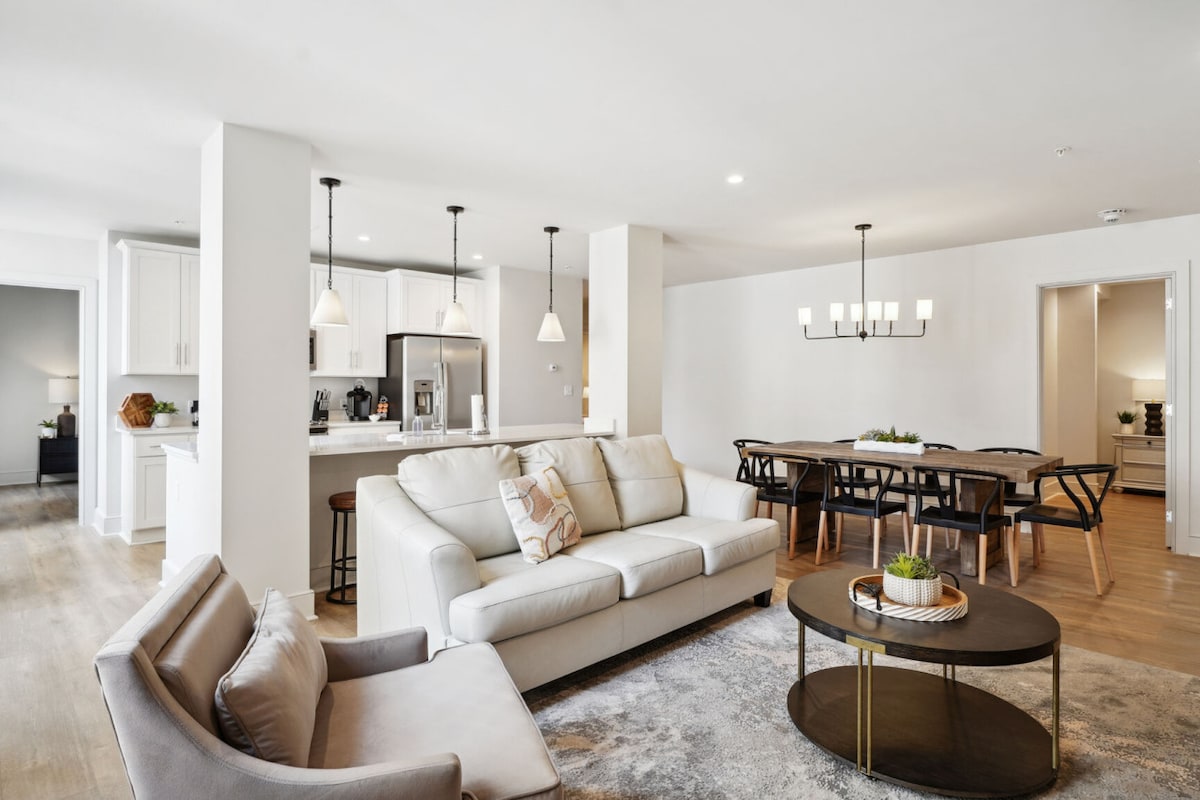 A bright and modern living space is configured with a spacious sofa and armchair. A round coffee table rests on a soft rug, while a dining table with stylish chairs is positioned nearby. The fully equipped kitchen is visible in the background, promoting a welcoming atmosphere.