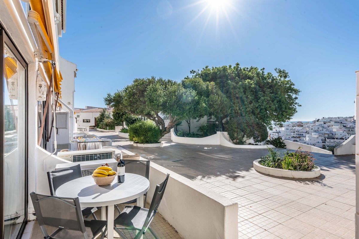 A sunlit terrace is showcased, featuring a round table surrounded by four chairs. Lush greenery adds a natural touch, while a large tree offers shade. The view includes the surrounding white structures of Albufeira, and a clear blue sky enhances the serene ambiance.