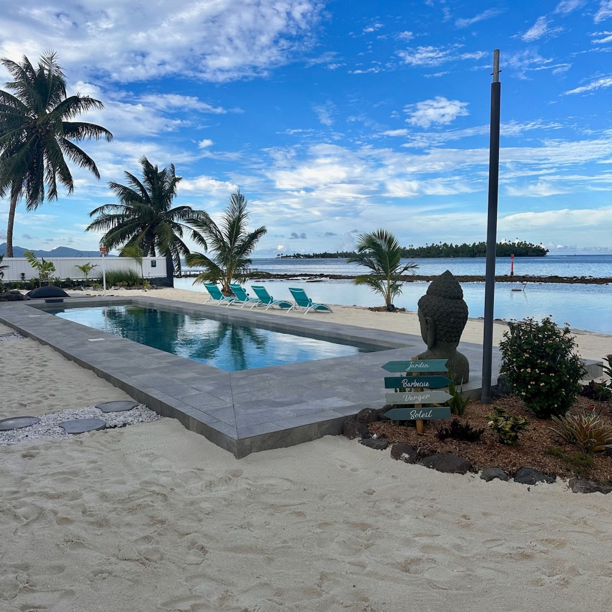 A serene swimming pool is surrounded by white sand, with lounge chairs positioned for relaxation. Tropical palm trees frame the area, and the lagoon reflects a clear blue sky. A decorative statue and signage add character to the landscape, enhancing the tranquil ambiance.