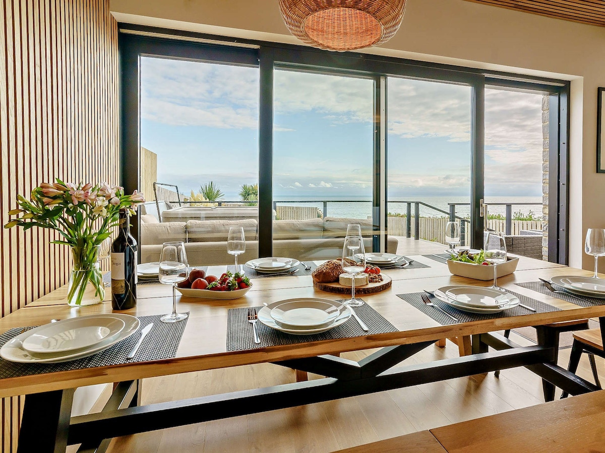 A modern dining area is framed by large glass doors revealing scenic sea views. The table is set with plates, wine glasses, and bowls of fresh food, while a bouquet of flowers adds a natural touch. Natural light enhances the inviting atmosphere.