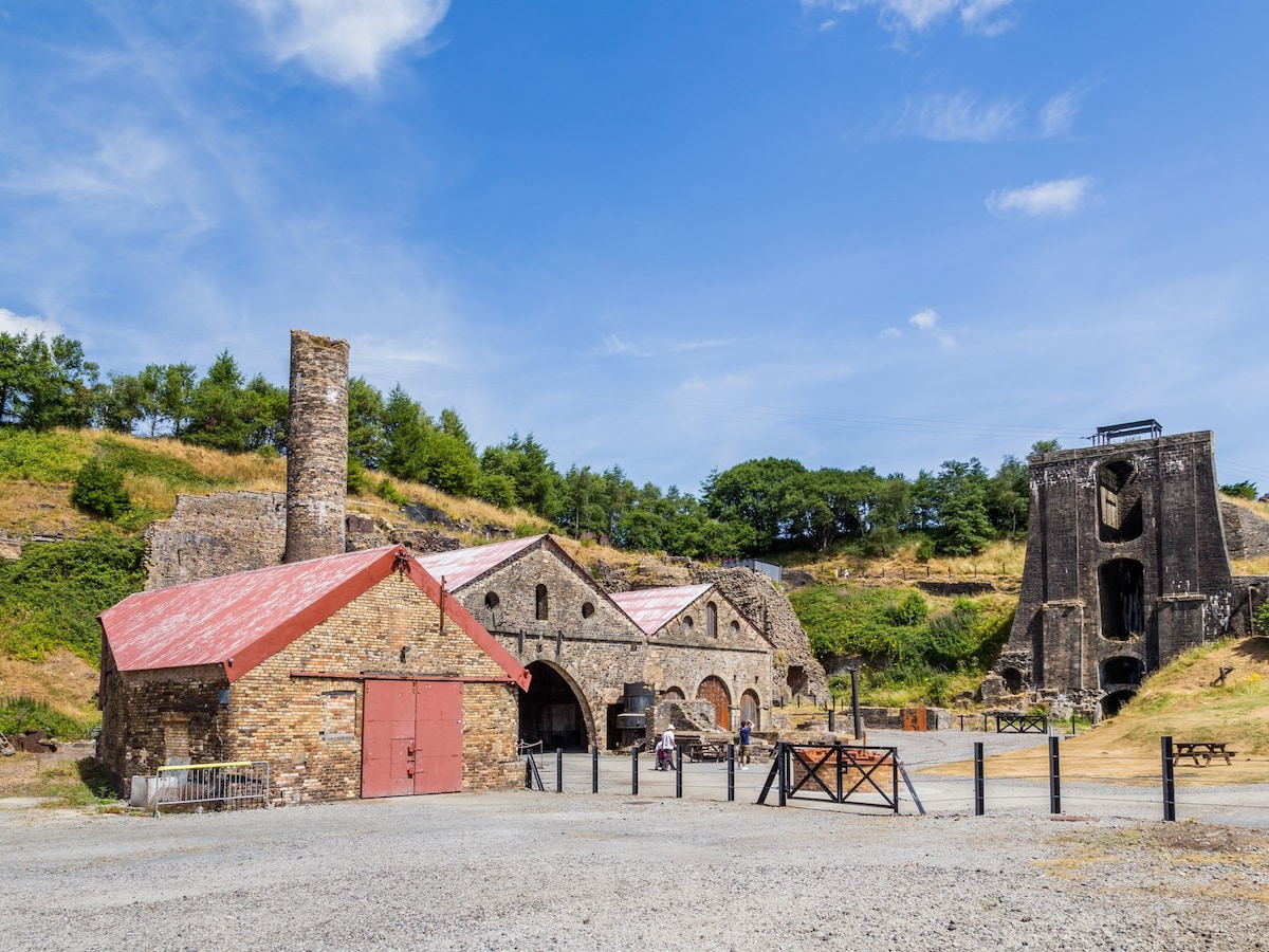 An industrial heritage site is depicted, featuring stone buildings with red roofs and large arched doorways. A chimney stack stands prominently, while remnants of a mine structure loom in the background. Surrounding greenery adds a natural contrast to the historical architecture.