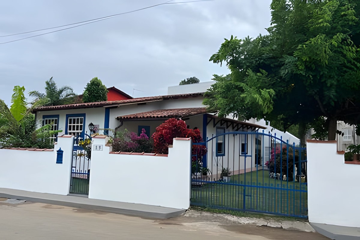 A single-story house features a colorful exterior with blue accents and a tiled roof. Lush greenery and vibrant flowering plants surround the entrance, which is marked by a decorative blue gate. A well-maintained garden enhances the welcoming appearance of the property.