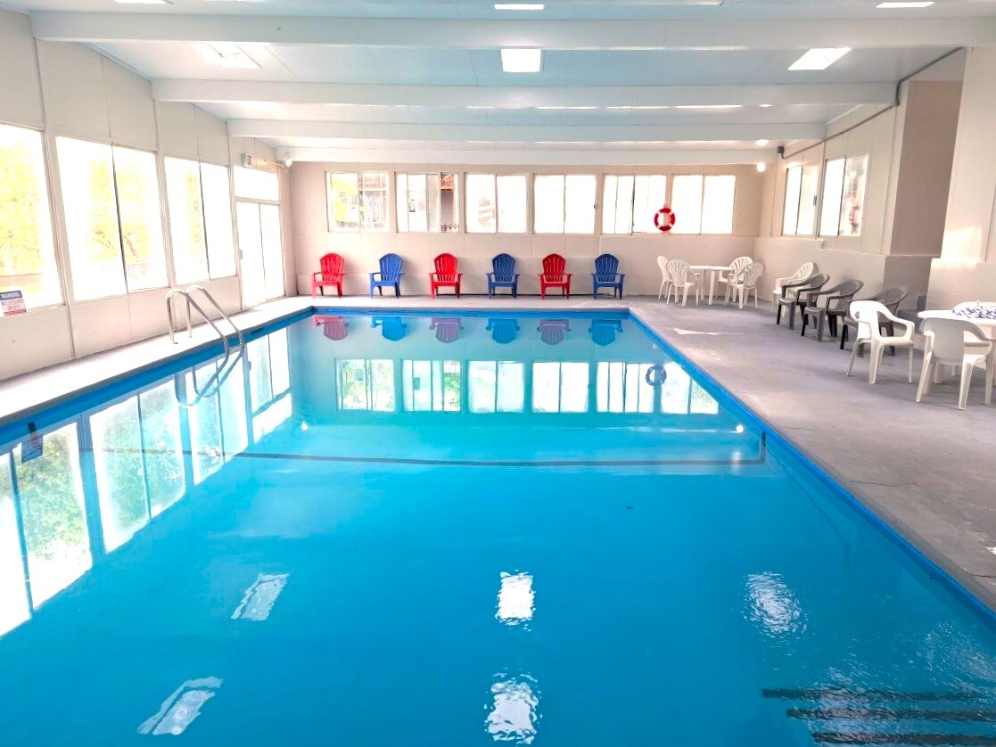 An indoor swimming pool is surrounded by large windows, allowing natural light to fill the space. Colorful lounge chairs line one side, with red and blue options contrasting against white plastic chairs. The clear blue water reflects the overhead lights, creating a calming atmosphere.