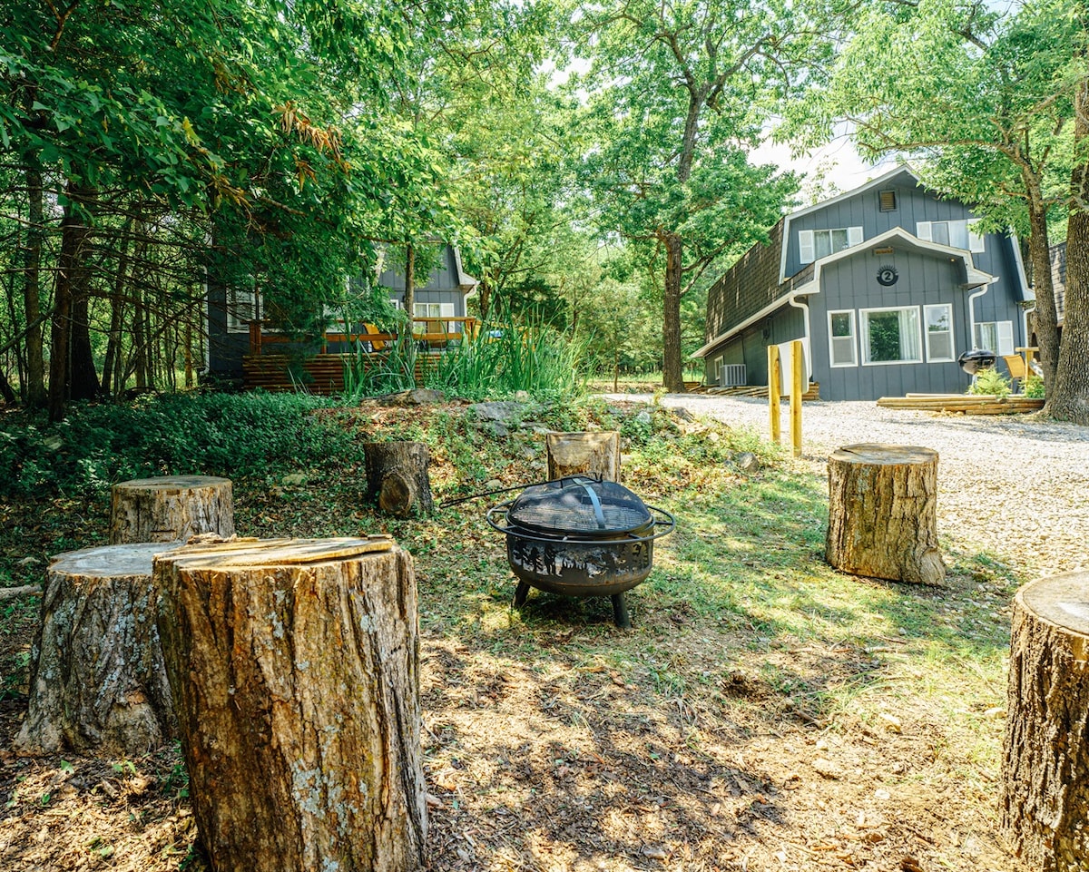 A fire pit is surrounded by tree stump seating, set amidst greenery and grass. The rustic black fire ring is visible, while the nearby cabins are partially obscured by trees, creating a natural and inviting outdoor space.