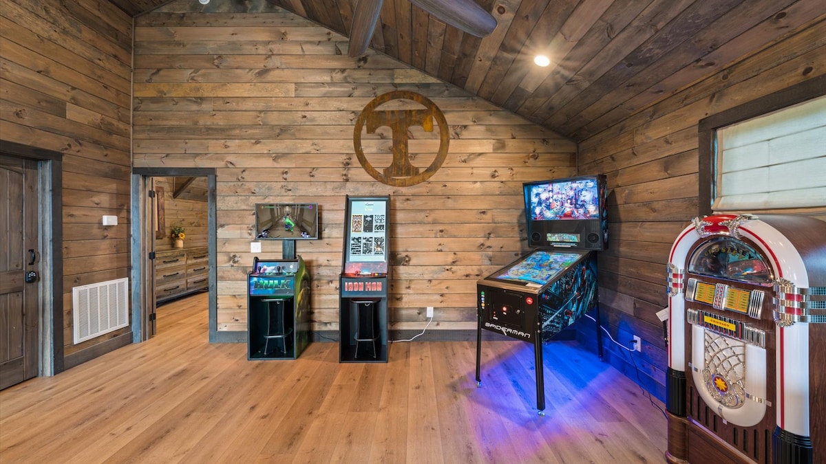 An open gaming loft is presented with wooden walls and a high ceiling. Various arcade machines are positioned along one wall, including a pinball machine and a jukebox, with soft lighting enhancing the inviting space. Ample floor area accommodates movement and play.