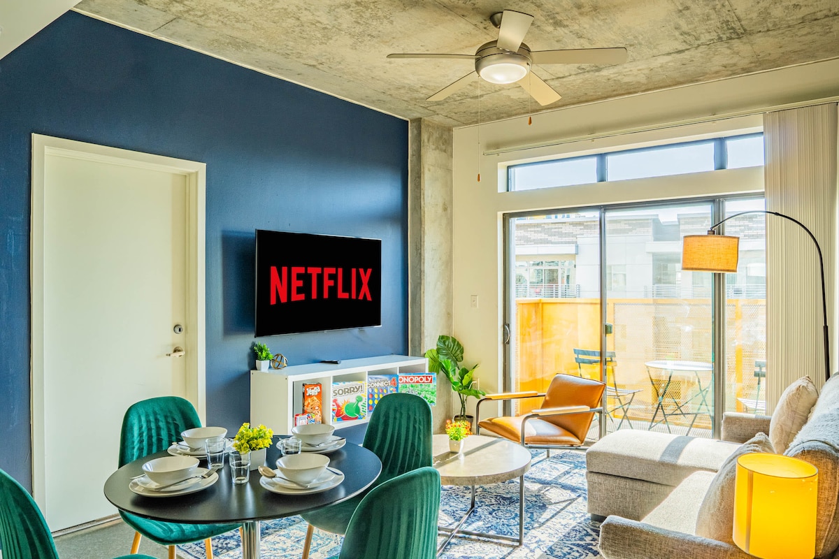 A modern living area features a sleek sofa, a round dining table set for four, and a 55" HDR Smart TV displaying the Netflix logo. Natural light fills the room through a large window, highlighting the tasteful decor and minimalist design.
