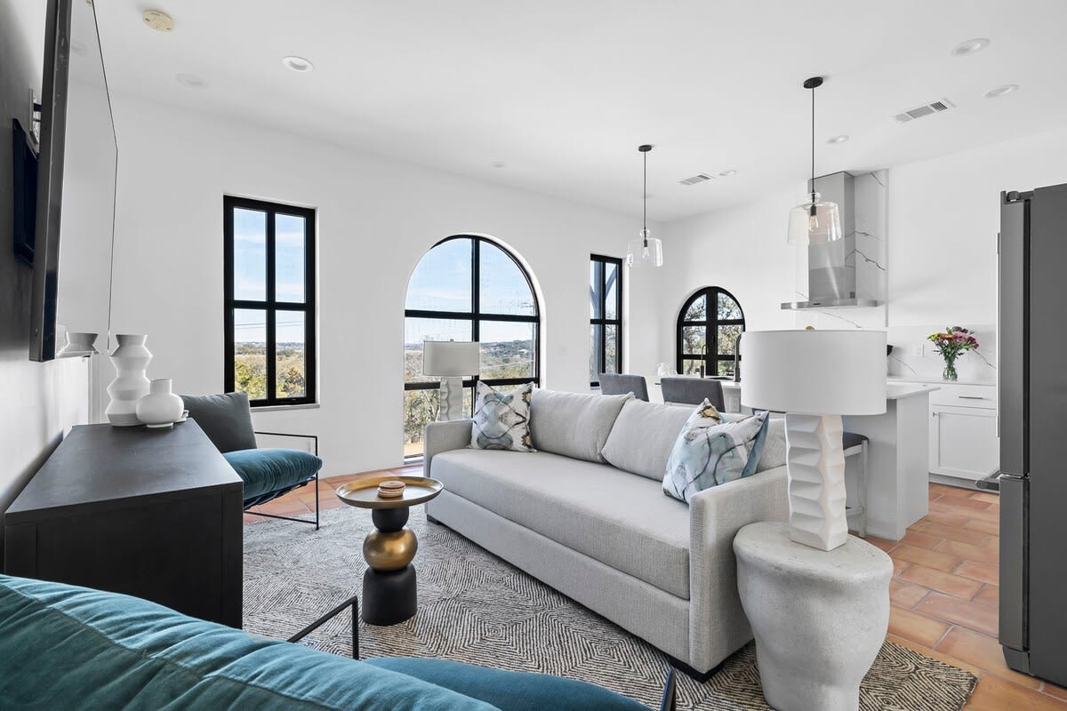 A bright living area features large arched windows that frame scenic valley views. A spacious light grey sofa, accented with soft blue and green pillows, invites relaxation. A round coffee table sits prominently, while a dining area and modern kitchen are visible in the background.