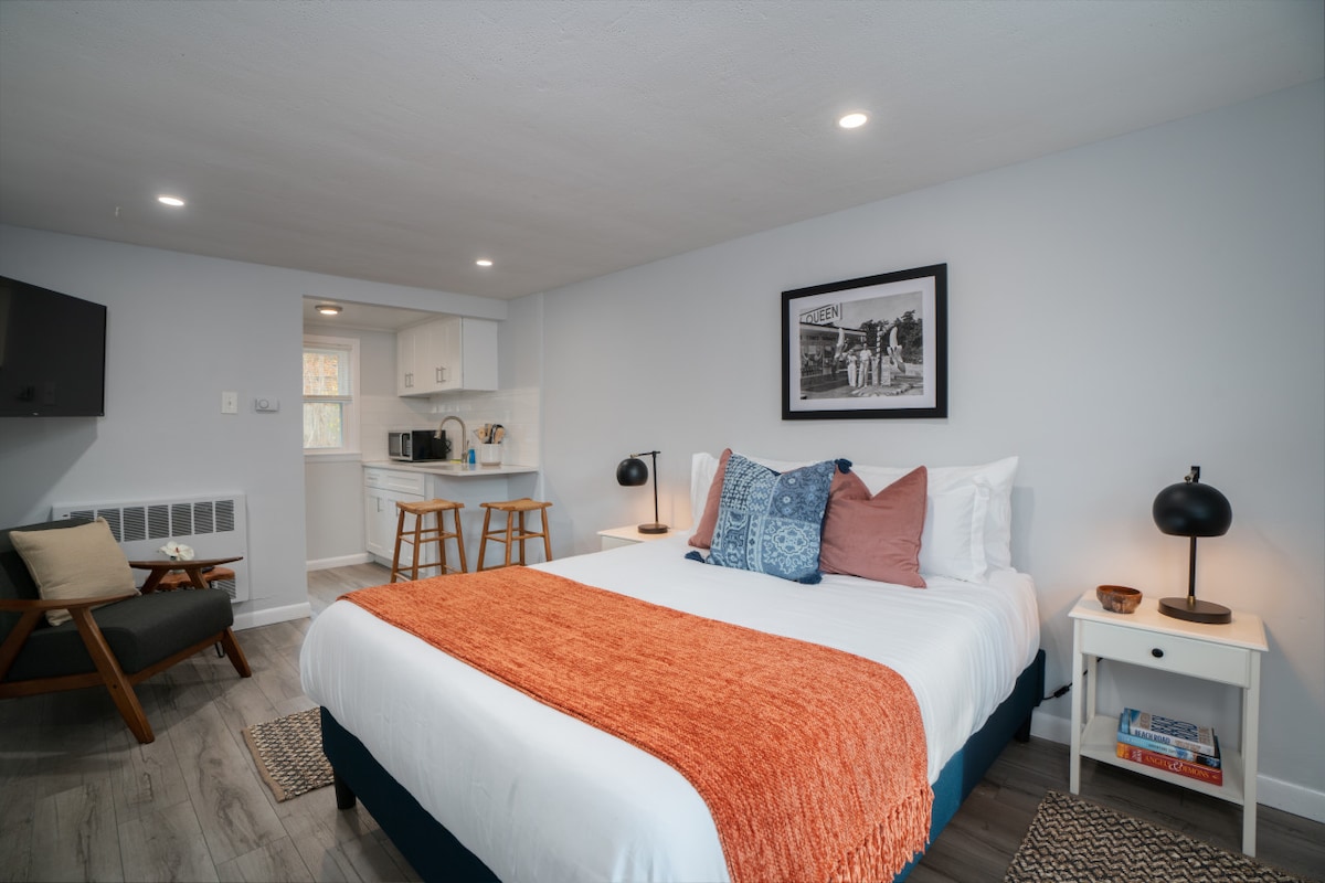 A bright studio apartment features a spacious queen bed with decorative pillows and an orange throw. A small kitchen area can be seen in the background, alongside a wall-mounted TV. Modern lighting fixtures provide soft illumination near the bed and a cozy armchair is positioned nearby.