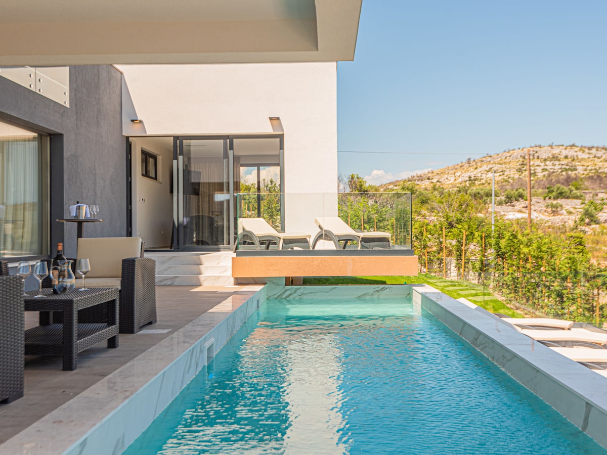 An outdoor space features a glistening swimming pool with an adjacent terrace furnished with lounge chairs. A seating area with a table and chairs is visible, providing a comfortable spot for relaxation. Lush greenery and a hillside can be seen in the background.