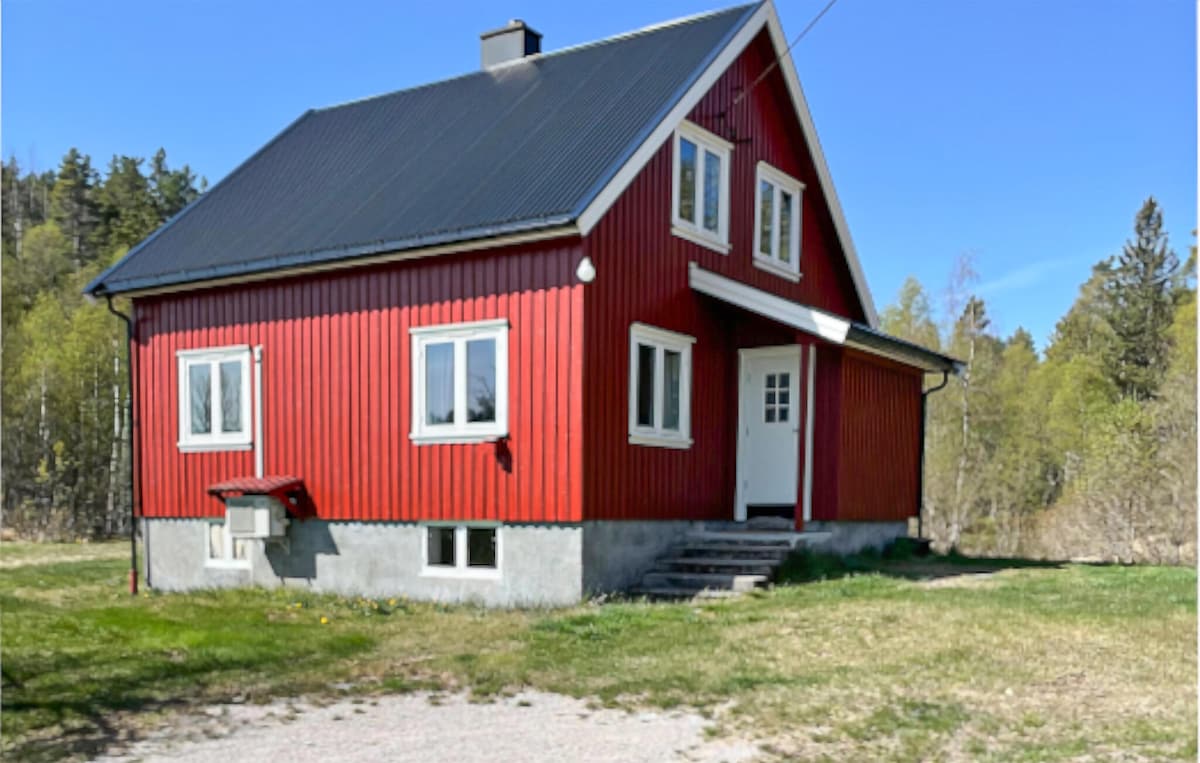 A red, two-story house with a sloped black roof is surrounded by greenery. Four windows adorn the front, providing natural light to the interior. A small set of steps leads to the front door, with a gravel pathway leading to the home.