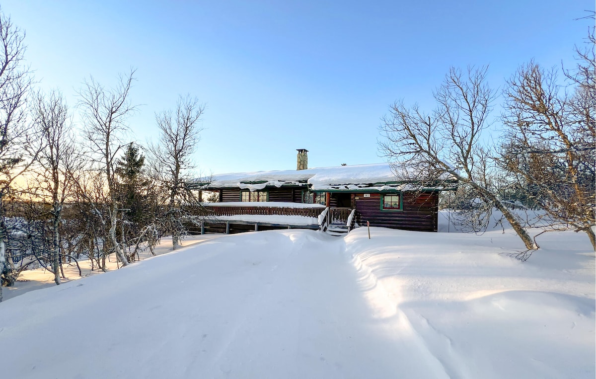 A cozy vacation home is surrounded by a blanket of fresh snow, with a pathway leading to the entrance. The structure features large windows, allowing natural light to fill the interior. Leafless trees frame the scene, contributing to the serene winter landscape.