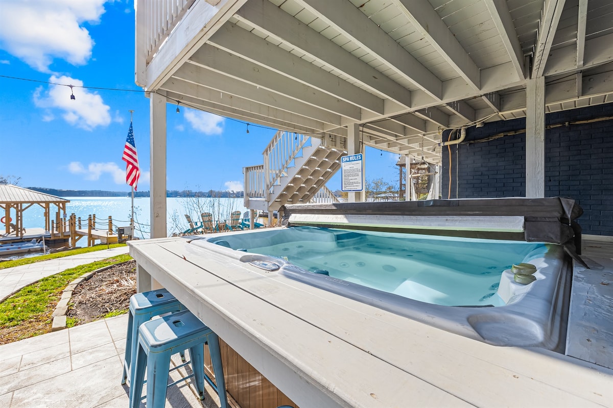 A hot tub sits on a wooden deck, surrounded by a serene landscape overlooking the lake. Bright blue skies reflect above, while a nearby American flag gently flutters in the breeze. Sturdy steps lead to a lower level, creating easy access to the outdoor space.