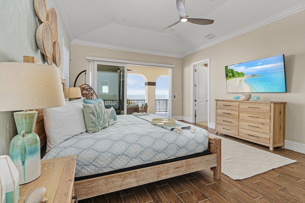 A spacious bedroom features a large bed with a light blue quilt and decorative pillows. Natural light enters through a glass door leading to a balcony, where scenic views are visible. A wooden dresser and bedside tables add warmth to the room, complemented by tasteful décor.