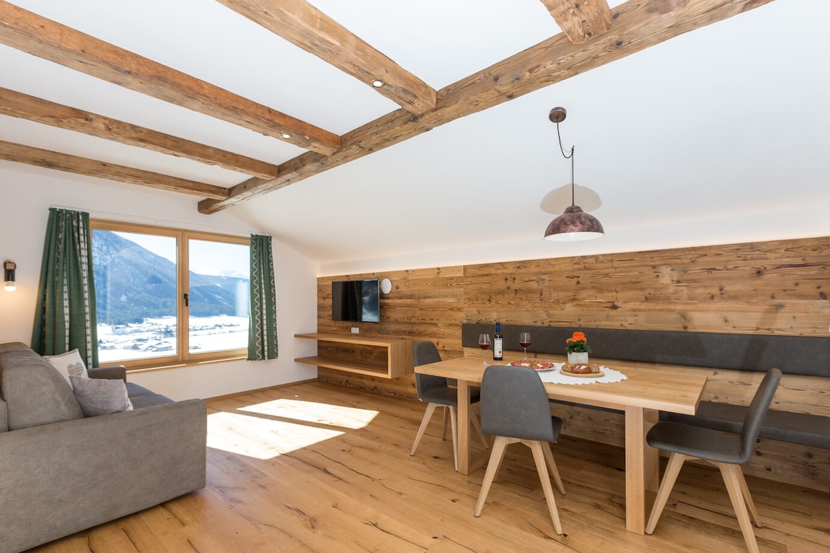 A spacious living area features wooden beams and a large window showcasing panoramic mountain views. A comfortable sofa is positioned adjacent to a dining table with two chairs, complemented by a TV mounted on the wall. Natural light brightens the warm wooden floors.