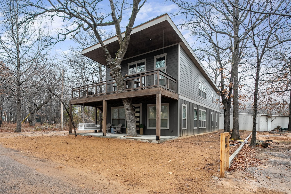 The exterior of a two-story home features a modern design with a wooden porch and large windows. Surrounding trees provide shade, while a spacious area in front is lined with gravel. The structure is inviting with a warm grey color and dark wood accents.