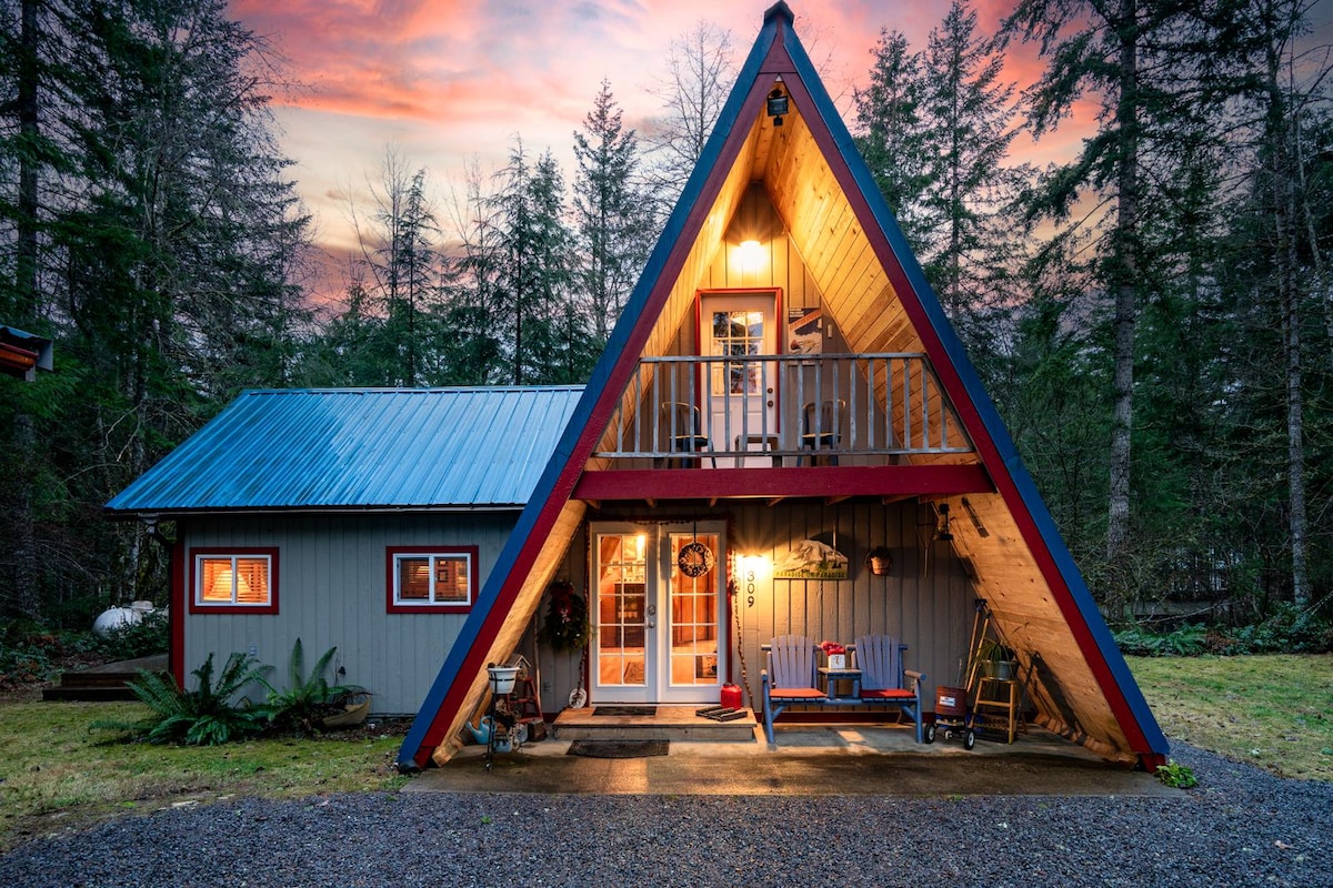 The A-frame cabin is set amidst tall trees, showcasing a sloped roof with vibrant blue and red detailing. A welcoming entrance features double doors, leading to a deck embellished with a small sitting area. The twilight sky casts a warm glow on the rustic exterior.