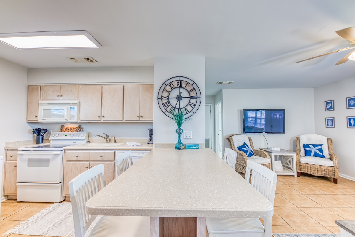 A spacious kitchen area features light beige cabinetry and a large countertop with seating for six. A clock decorates the wall above the countertop, while appliances are arranged for convenience. A cozy seating area with a television can be seen in the background.