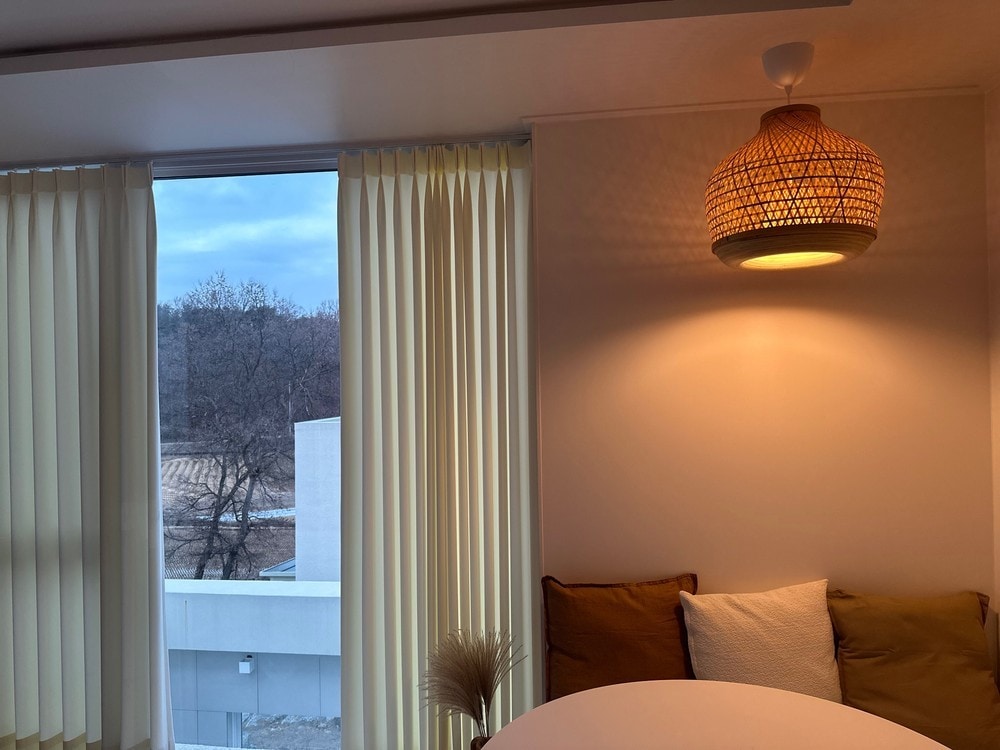A cozy corner features soft, warm lighting from a woven pendant lamp, accentuating a small round table surrounded by plush cushions. Sheer curtains frame the window, revealing a serene view of the outdoors, blending the interior and exterior spaces.
