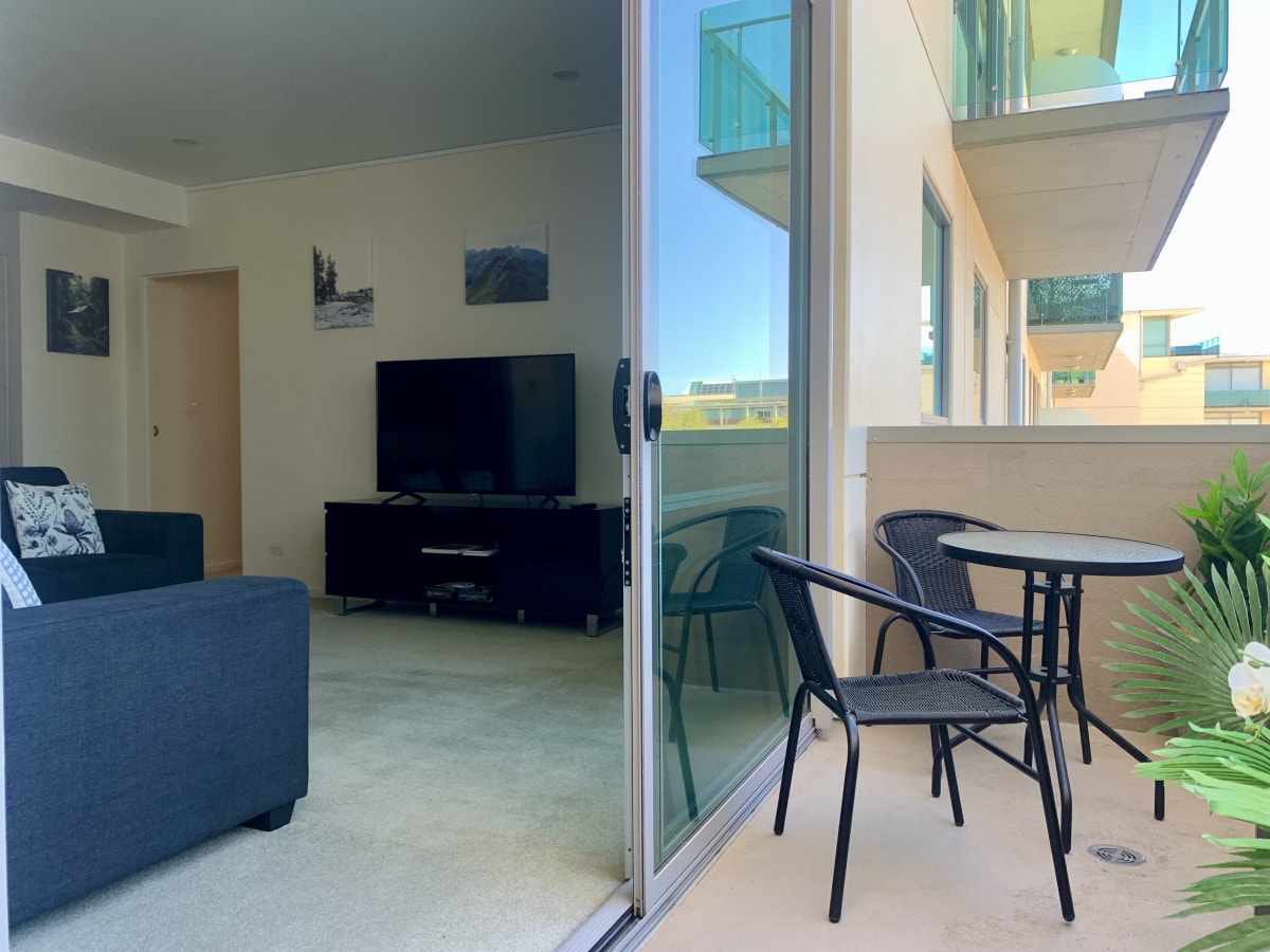 A spacious balcony is featured, complete with a small table and two chairs. A living area is visible through large sliding glass doors, showcasing a television and a cozy sofa, with soft carpeting and decorative wall art in the background.