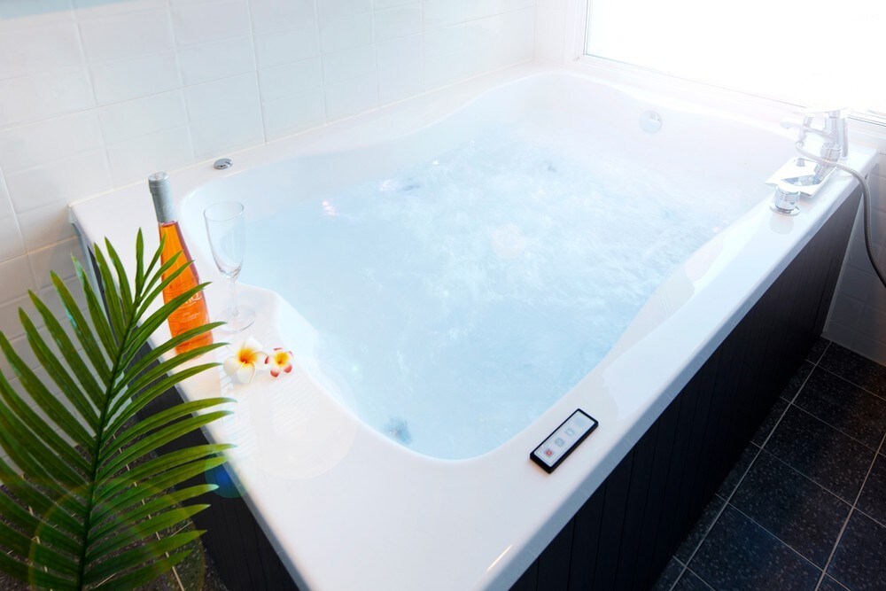 A modern whirlpool bath is set against a bright window, creating a serene bathing space. Adjacent to the tub, a glass of sparkling wine and a bottle rest on the ledge, complemented by a sprig of greenery and a few tropical flowers.