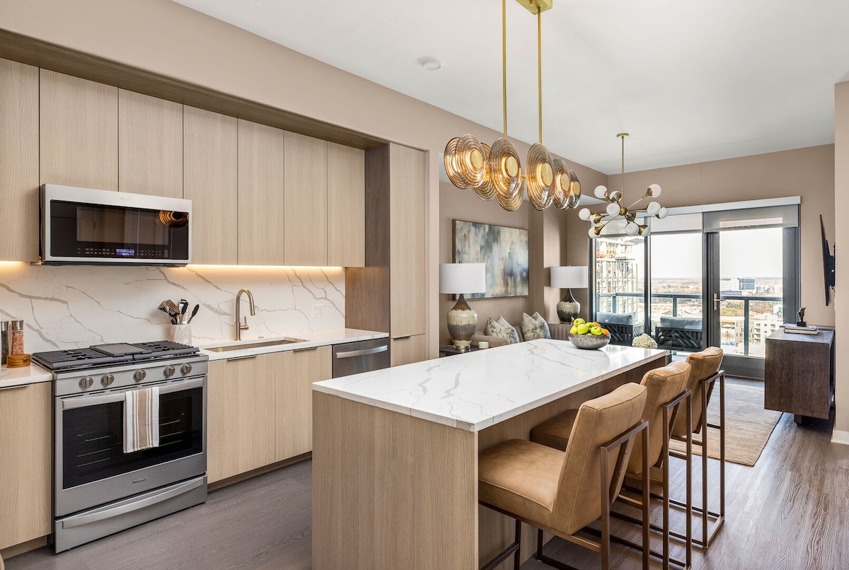The kitchen features sleek wooden cabinetry and a marble-topped island with seating for four. High-end appliances, including a gas stove and microwave, are visible. A dining area is adorned with modern light fixtures, and large windows offer a view of the outside balcony.
