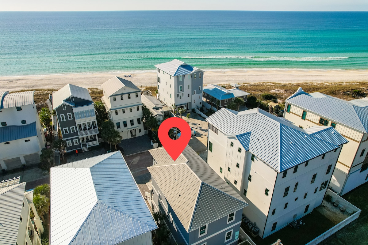 An aerial view captures the property surrounded by vibrant coastal scenery, highlighting the proximity to the beach. The building features a metal roof and distinct architectural details, with sandy shorelines and shimmering waters visible in the background.