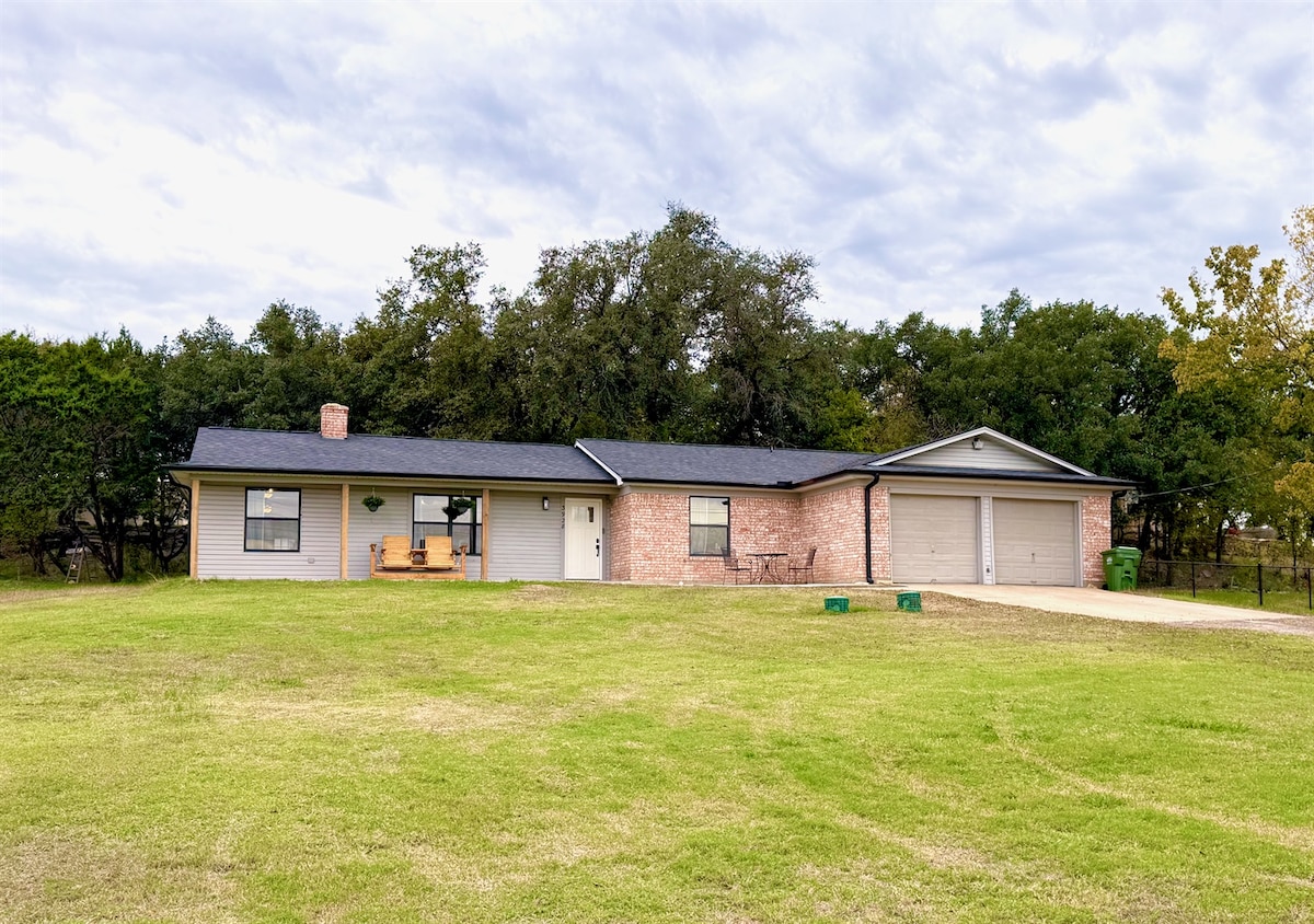 A spacious hilltop home is set on a green lawn, featuring a front porch with a swing. Two garage doors are visible, and the exterior is designed with a mix of brick and siding under a cloudy sky.