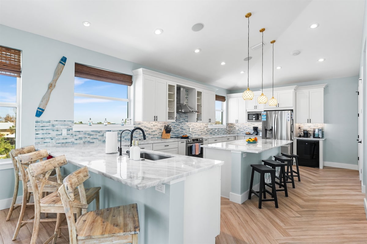A spacious kitchen is highlighted by a large island with bar seating for six. Modern appliances are integrated into white cabinetry, and a light-colored backsplash enhances the design. Natural light floods the area through large windows, providing views of the outdoors.