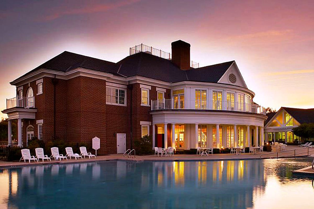 A large, elegant building is positioned near a swimming pool, reflecting the warm colors of the sunset. Expansive windows and a combination of brick and siding highlight the architecture. Lounge chairs line the poolside, creating an inviting outdoor space for relaxation.
