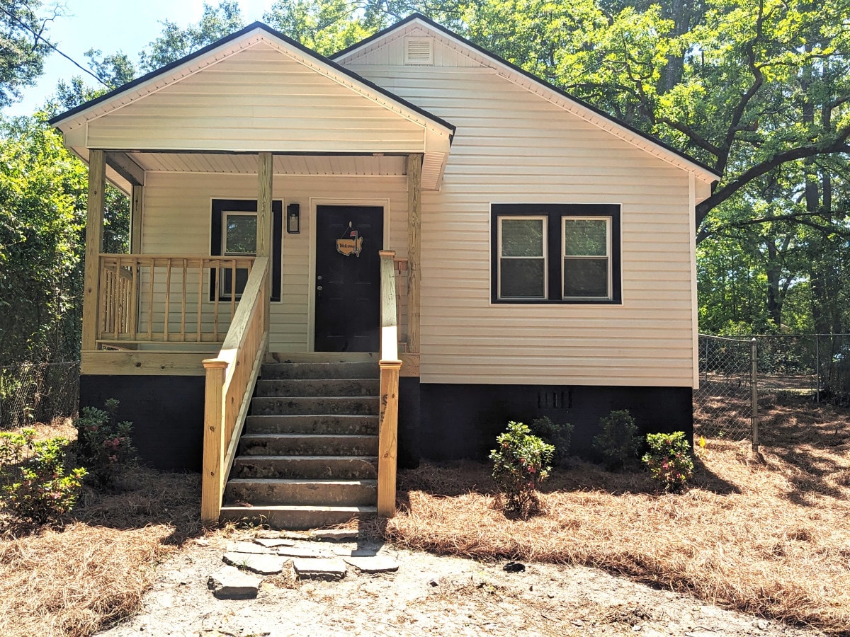 The exterior of the cottage showcases a welcoming front porch with steps leading up to the entrance. A simple black door is framed by two windows on either side. Surrounding vegetation includes small shrubs, and the natural landscaping features pine needles covering the ground.