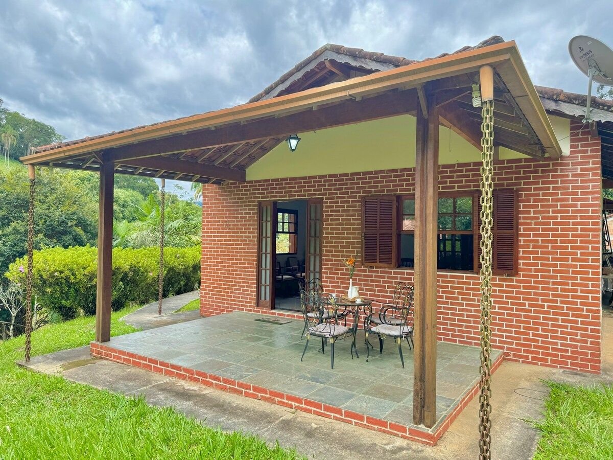 A covered patio area features a stone floor and a set of metal chairs surrounding a small table. The brick exterior of the house contrasts with the lush green surroundings, offering a peaceful setting with nature visible in the background.