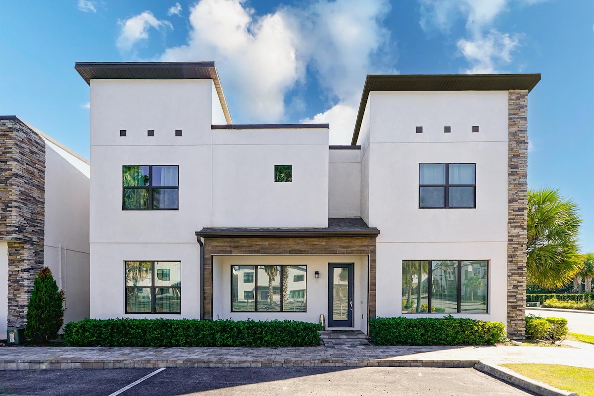 A modern two-story villa with a clean white facade and contrasting stone accents. Large windows allow natural light to fill the interior, while a well-kept flower bed is presented in front. The building offers a welcoming appearance with an inviting entrance and well-defined architectural features.