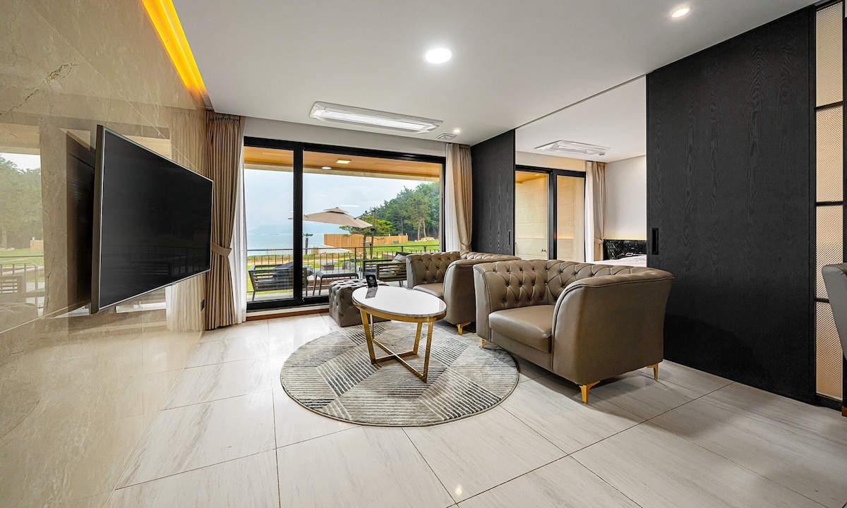 The living area features comfortable seating arrangements with a sleek coffee table in the center. Large windows offer expansive views of the outdoors, while natural light highlights the marble flooring. A flat-screen TV is mounted on the wall, complementing the modern decor.