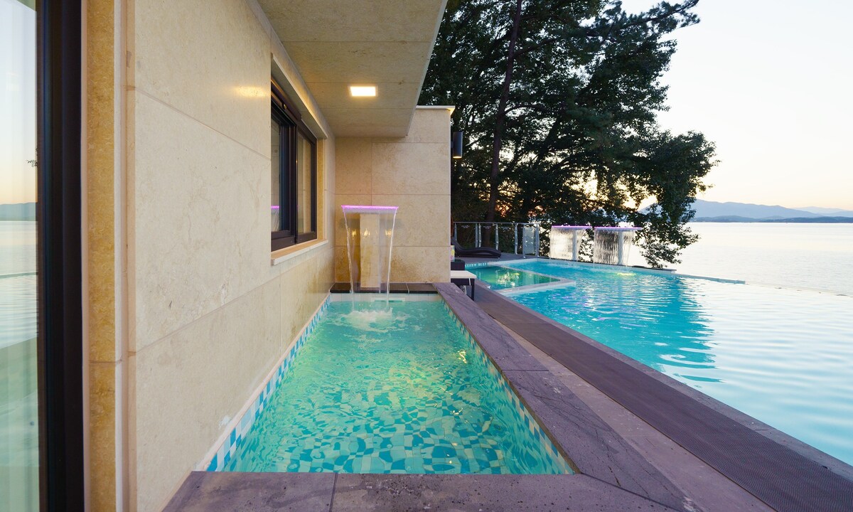 A tranquil infinity pool is shown beside a sleek stone structure, with a gentle waterfall feature. The water reflects soft hues from the setting sun, surrounded by lush trees and a stunning view of the ocean in the background.