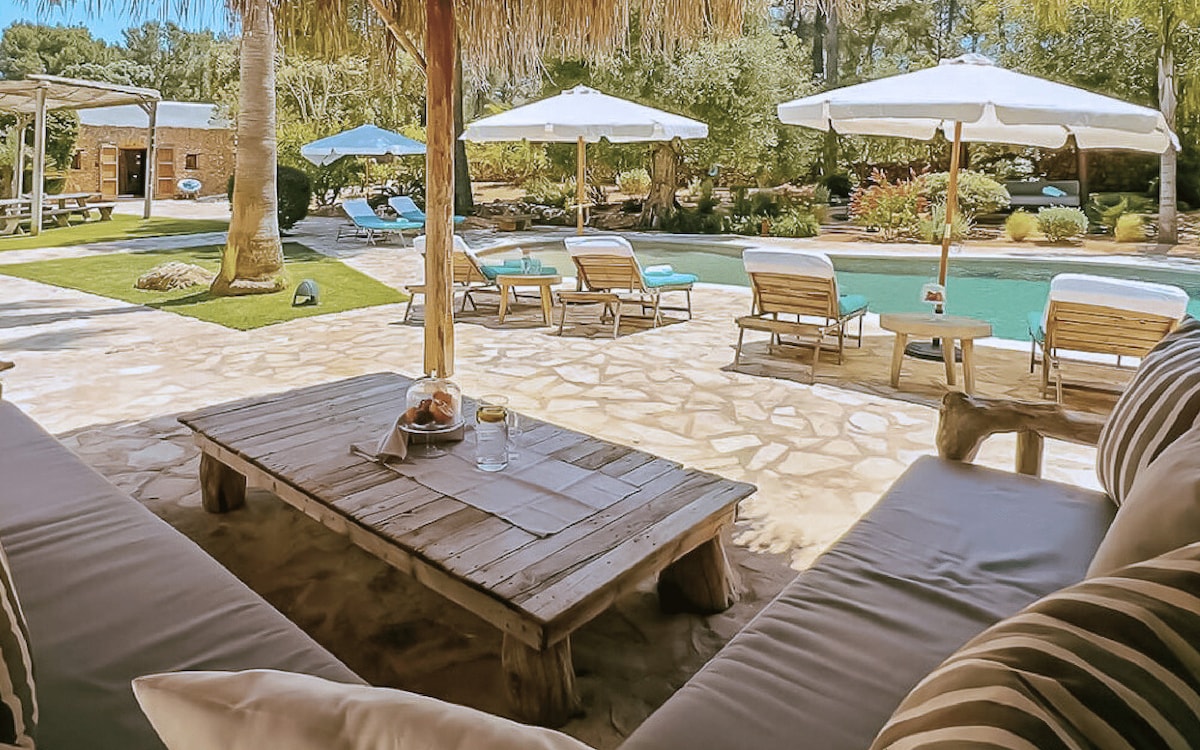 A shaded outdoor lounge area is presented, featuring a rustic wooden table with a breakfast setup. Sun loungers and umbrellas are visible around the inviting pool, surrounded by lush greenery and an overall tranquil atmosphere, perfect for relaxation in the Ibiza sun.