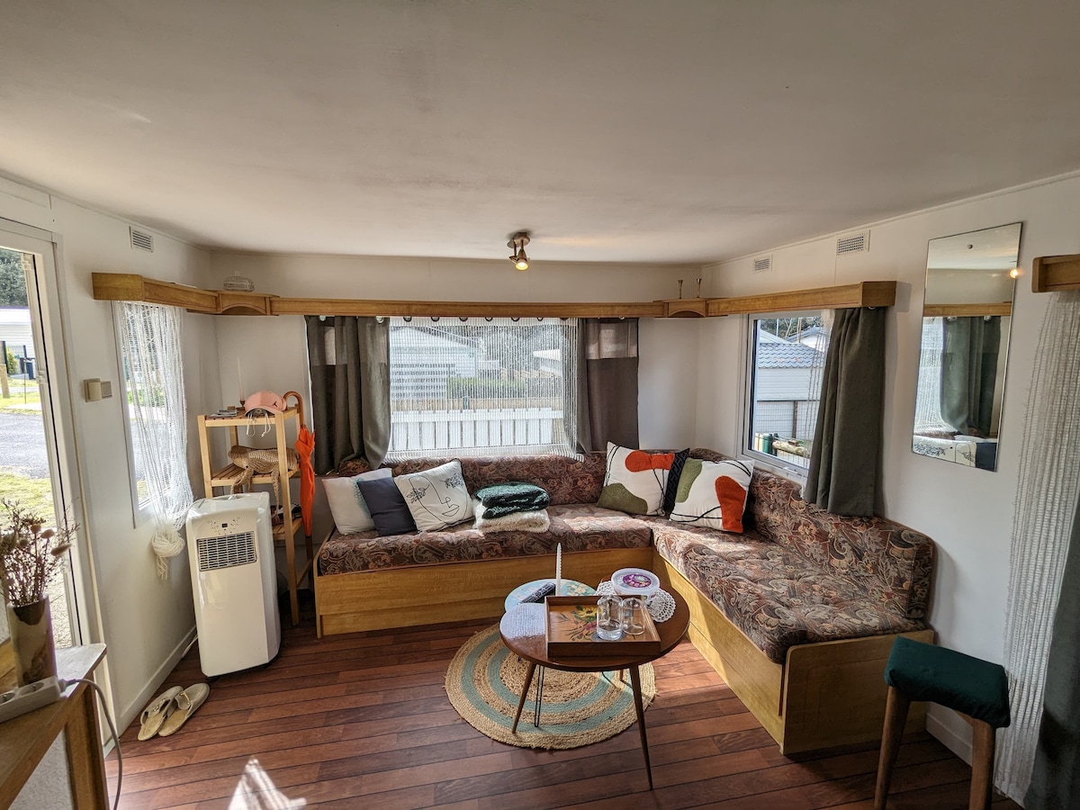 A lounge area features a comfortable L-shaped sofa adorned with colorful cushions, inviting relaxation. A round coffee table sits at the center, surrounded by natural light from large windows with sheer curtains, providing a view of the outside. A small shelf is positioned in the corner.